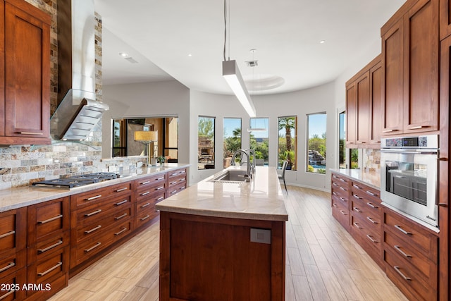 kitchen featuring a center island with sink, a sink, tasteful backsplash, appliances with stainless steel finishes, and wall chimney exhaust hood