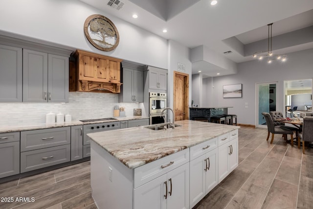 kitchen with backsplash, gray cabinetry, double oven, light stone counters, and a sink