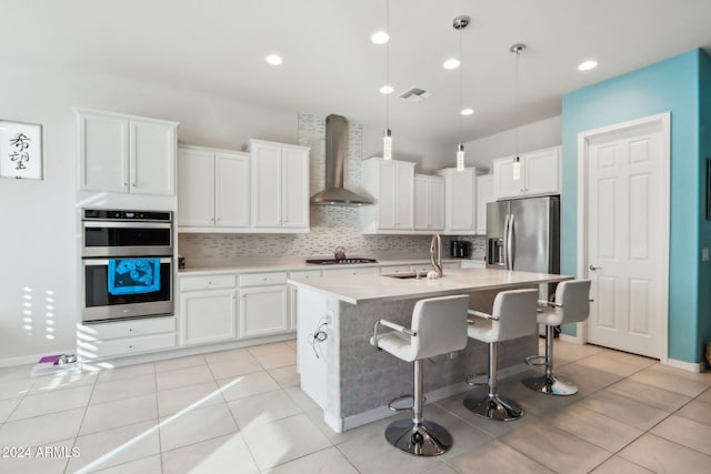 kitchen with white cabinetry, wall chimney exhaust hood, decorative light fixtures, a center island with sink, and appliances with stainless steel finishes