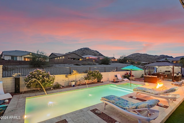 pool at dusk featuring pool water feature, a mountain view, a gazebo, and an outdoor fire pit