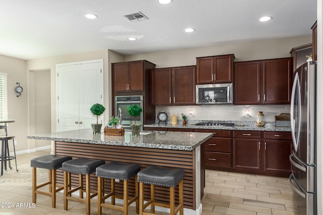 kitchen featuring a kitchen island with sink, backsplash, appliances with stainless steel finishes, a kitchen breakfast bar, and light stone countertops