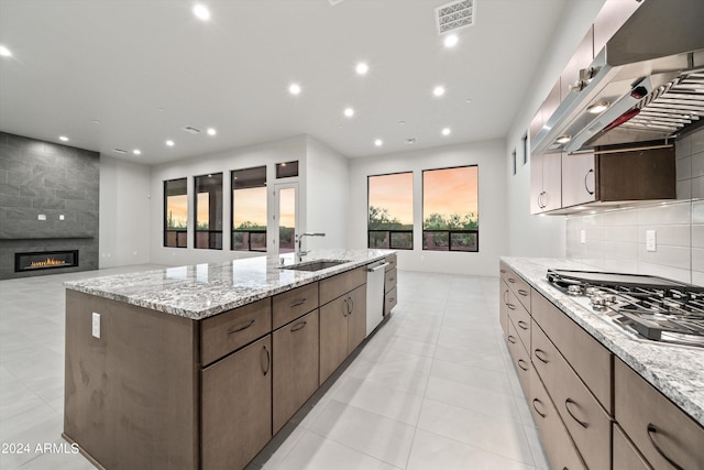 kitchen featuring a kitchen island with sink, a large fireplace, range hood, sink, and appliances with stainless steel finishes