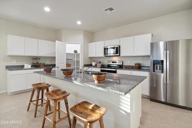 kitchen with white cabinets, stainless steel appliances, dark stone countertops, and a kitchen island with sink