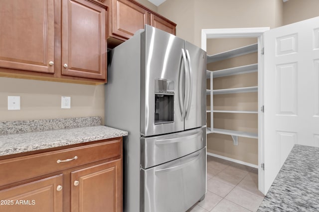 kitchen with light tile patterned floors, brown cabinetry, stainless steel fridge, and light stone countertops