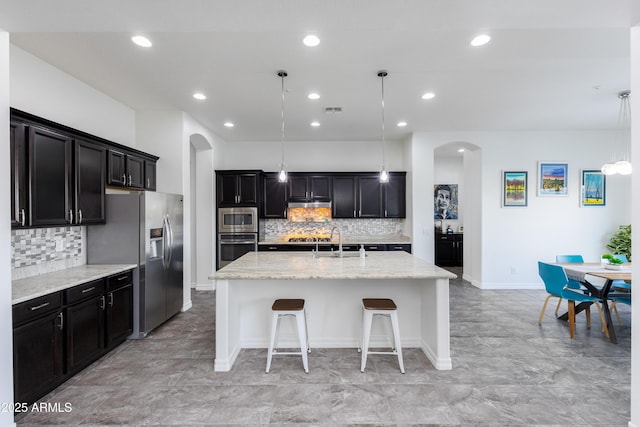 kitchen with arched walkways, light stone counters, decorative light fixtures, appliances with stainless steel finishes, and a center island with sink