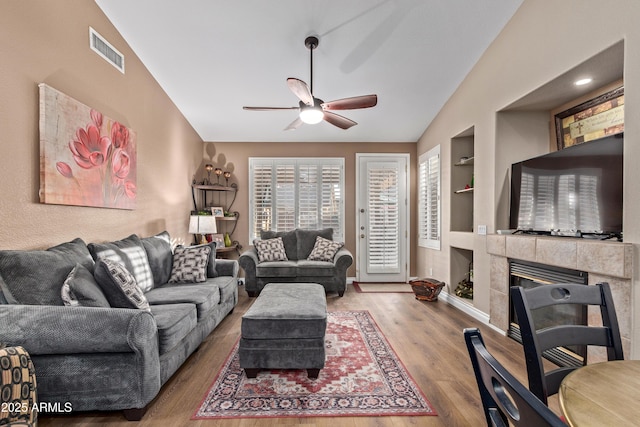 living room with built in shelves, ceiling fan, a tile fireplace, hardwood / wood-style flooring, and lofted ceiling