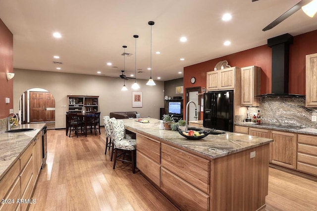 kitchen with a center island with sink, light hardwood / wood-style flooring, light stone countertops, wall chimney exhaust hood, and ceiling fan