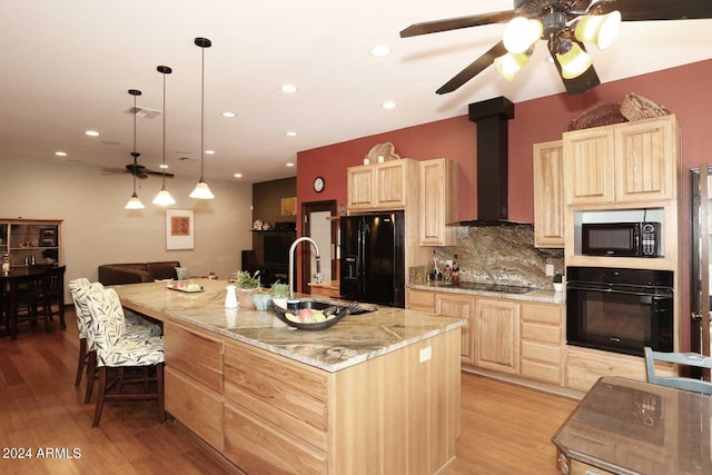 kitchen featuring black appliances, light wood-type flooring, an island with sink, light stone counters, and wall chimney exhaust hood