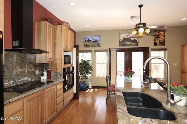 kitchen featuring sink, light stone countertops, black appliances, wall chimney range hood, and tasteful backsplash