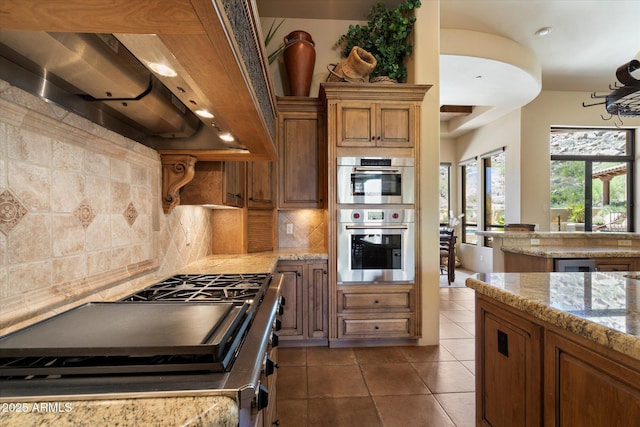 7731 E Soaring Eagle Way, Scottsdale, AZ 85266 - kitchen featuring ventilation hood, stainless steel double oven, backsplash, brown cabinets, and tile patterned flooring
