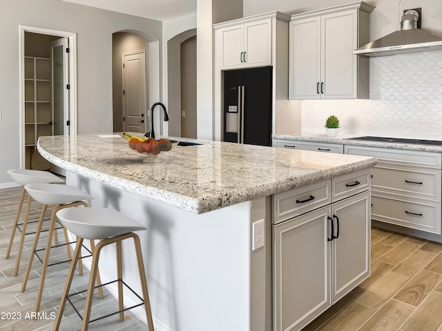 kitchen featuring backsplash, light hardwood / wood-style flooring, black appliances, wall chimney range hood, and a kitchen island with sink