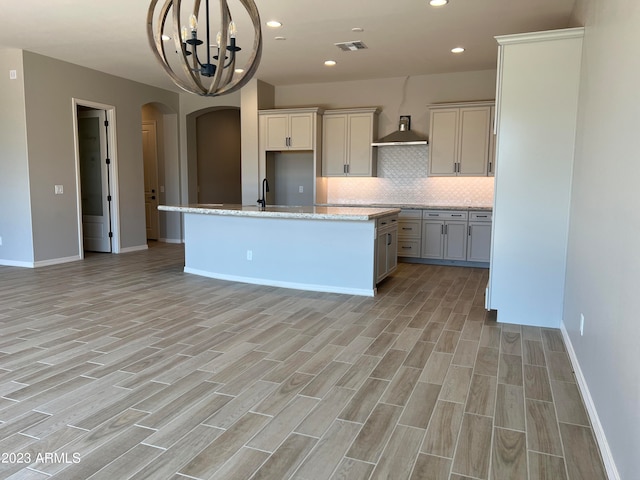 kitchen with light stone counters, wall chimney range hood, backsplash, a chandelier, and a kitchen island with sink