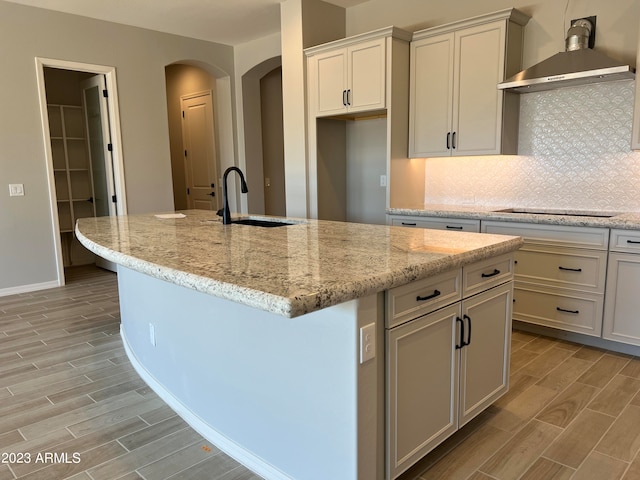 kitchen featuring wall chimney exhaust hood, a kitchen island with sink, light stone countertops, sink, and tasteful backsplash