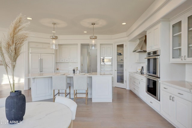 kitchen with appliances with stainless steel finishes, a center island with sink, pendant lighting, and wall chimney exhaust hood