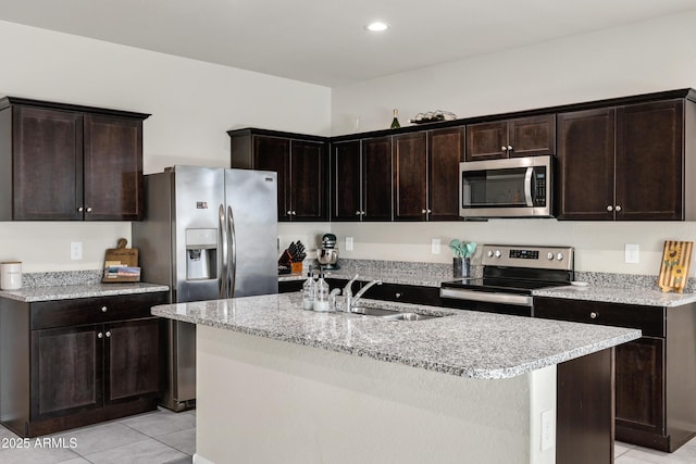 kitchen featuring dark brown cabinetry, a center island with sink, light tile patterned floors, appliances with stainless steel finishes, and a sink