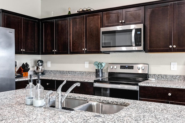 kitchen with dark brown cabinetry, light stone countertops, stainless steel appliances, and a sink