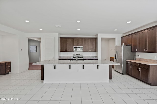 kitchen featuring dark brown cabinetry, an island with sink, appliances with stainless steel finishes, light stone countertops, and a kitchen bar