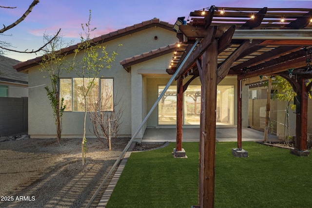 back of house at dusk with stucco siding, a lawn, a patio area, fence, and a tiled roof