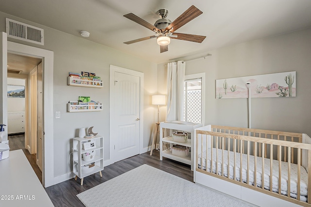 bedroom featuring visible vents, baseboards, a crib, a ceiling fan, and dark wood finished floors