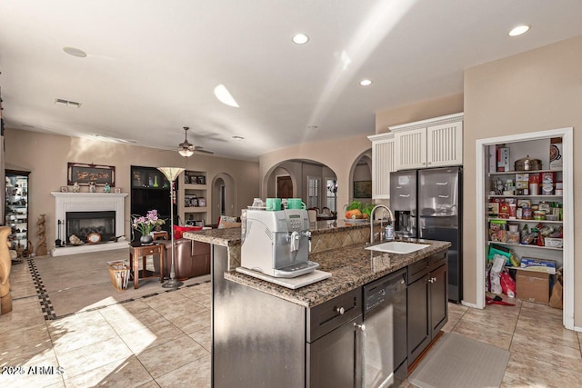 kitchen with dishwashing machine, a fireplace with raised hearth, a kitchen island with sink, arched walkways, and a sink