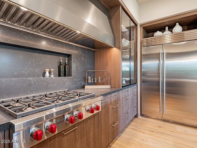 kitchen with light wood finished floors, tasteful backsplash, wall chimney exhaust hood, appliances with stainless steel finishes, and brown cabinets