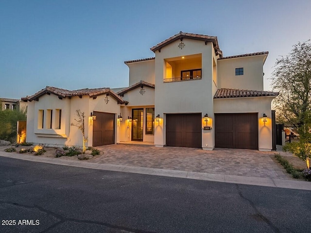 mediterranean / spanish house with a balcony, a garage, a tiled roof, decorative driveway, and stucco siding