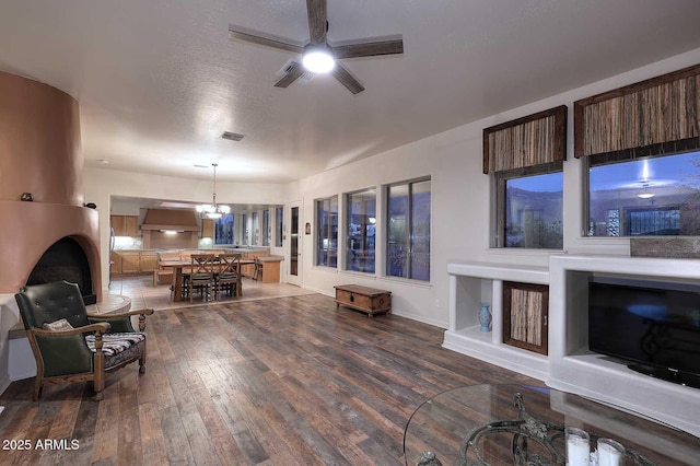 living area with a textured ceiling, ceiling fan with notable chandelier, visible vents, baseboards, and dark wood-style floors