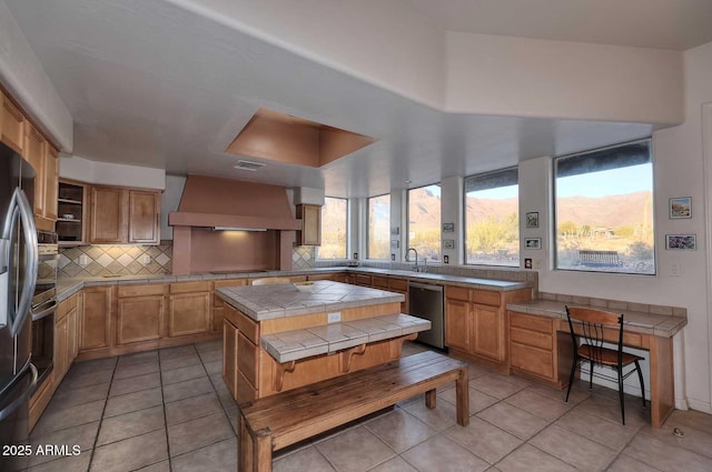 kitchen featuring wall chimney range hood, appliances with stainless steel finishes, tile counters, a center island, and tasteful backsplash