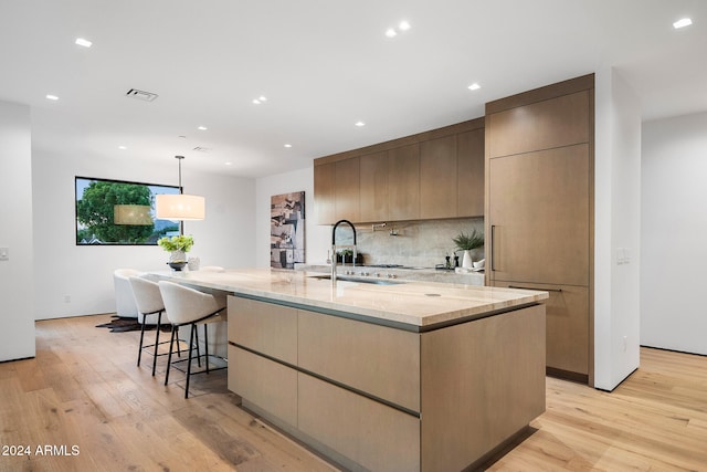 kitchen with light wood-type flooring, light stone counters, sink, a large island with sink, and hanging light fixtures