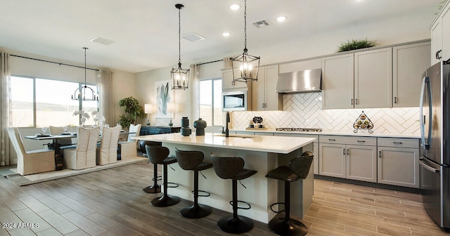 kitchen with decorative light fixtures, appliances with stainless steel finishes, backsplash, a center island with sink, and wall chimney range hood