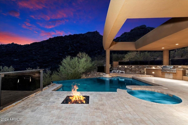 pool at dusk featuring a patio, a mountain view, an outdoor fire pit, an outdoor kitchen, and an in ground hot tub