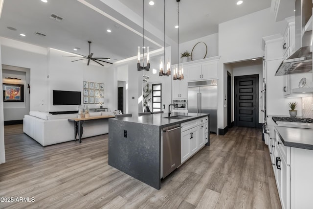 kitchen with white cabinetry, hanging light fixtures, a center island with sink, stainless steel appliances, and wall chimney range hood