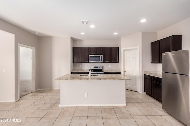 kitchen featuring dark brown cabinetry, light stone counters, appliances with stainless steel finishes, light tile patterned floors, and a kitchen island with sink