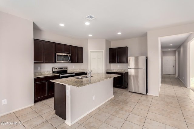 kitchen with dark brown cabinetry, sink, light stone counters, appliances with stainless steel finishes, and a kitchen island with sink