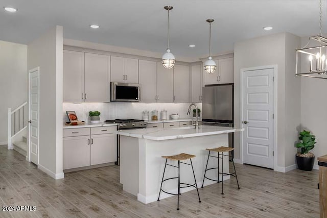 kitchen with tasteful backsplash, light wood-type flooring, an island with sink, stainless steel appliances, and pendant lighting