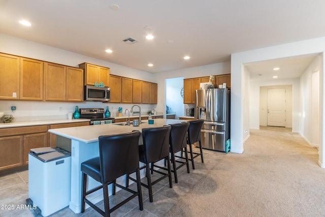 kitchen featuring appliances with stainless steel finishes, brown cabinetry, a kitchen bar, a center island with sink, and light countertops
