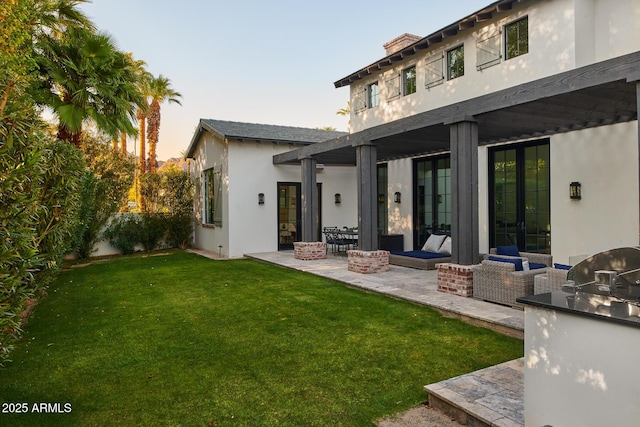 back house at dusk featuring exterior kitchen, an outdoor living space with a fire pit, a patio, and a lawn