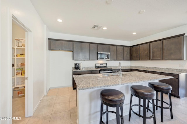 kitchen featuring sink, stainless steel appliances, light stone countertops, dark brown cabinets, and a center island with sink