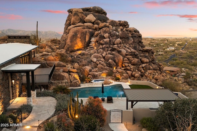 pool at dusk with a warm lit fireplace, an outdoor pool, a patio, a gate, and a mountain view