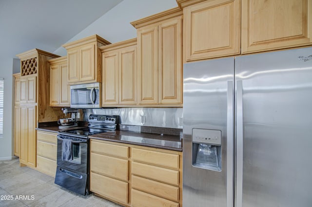 kitchen featuring dark stone countertops, appliances with stainless steel finishes, light brown cabinetry, and vaulted ceiling