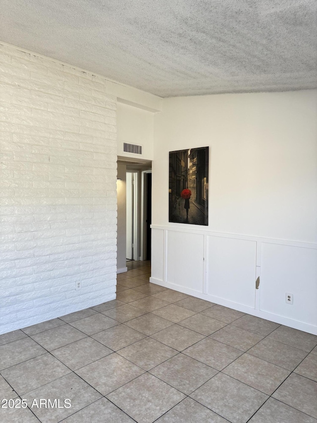 tiled empty room with visible vents, a textured ceiling, brick wall, and wainscoting