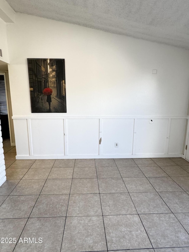 unfurnished room with a textured ceiling, wainscoting, a decorative wall, light tile patterned floors, and vaulted ceiling