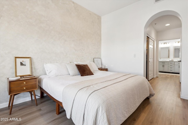 bedroom with baseboards, arched walkways, ensuite bath, and wood finished floors