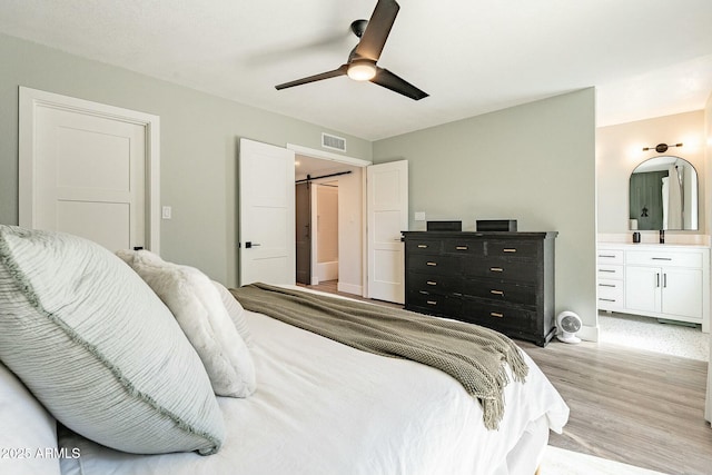 bedroom with a barn door, connected bathroom, a ceiling fan, visible vents, and light wood-style floors