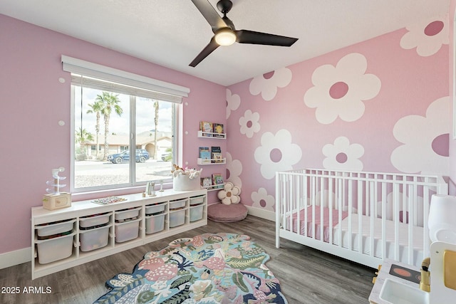 bedroom featuring a crib, baseboards, ceiling fan, an accent wall, and wood finished floors