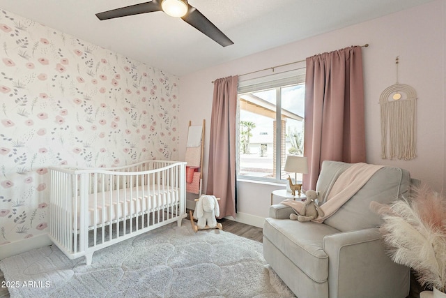 bedroom with a crib, wallpapered walls, baseboards, ceiling fan, and wood finished floors