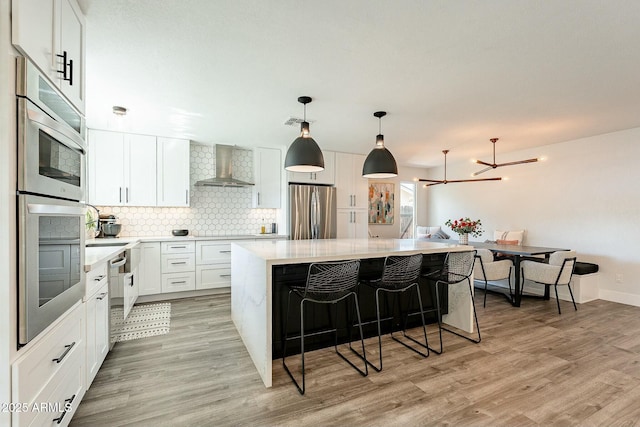 kitchen featuring a breakfast bar area, stainless steel appliances, backsplash, light wood-style floors, and wall chimney range hood