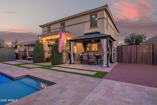 back of house at dusk with stucco siding, a gate, fence, a gazebo, and a patio area