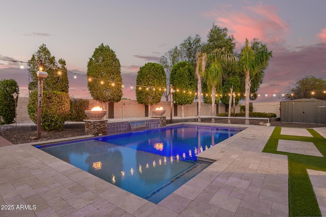 pool at dusk with a patio, a shed, a fenced backyard, and an outbuilding