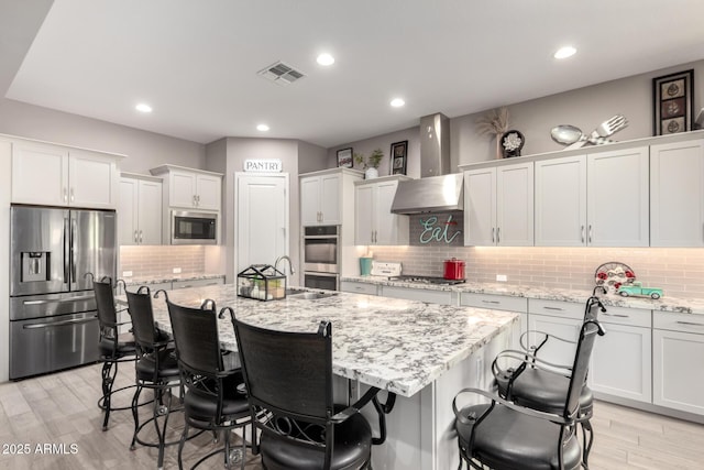 kitchen with appliances with stainless steel finishes, white cabinetry, light stone countertops, a center island with sink, and wall chimney exhaust hood